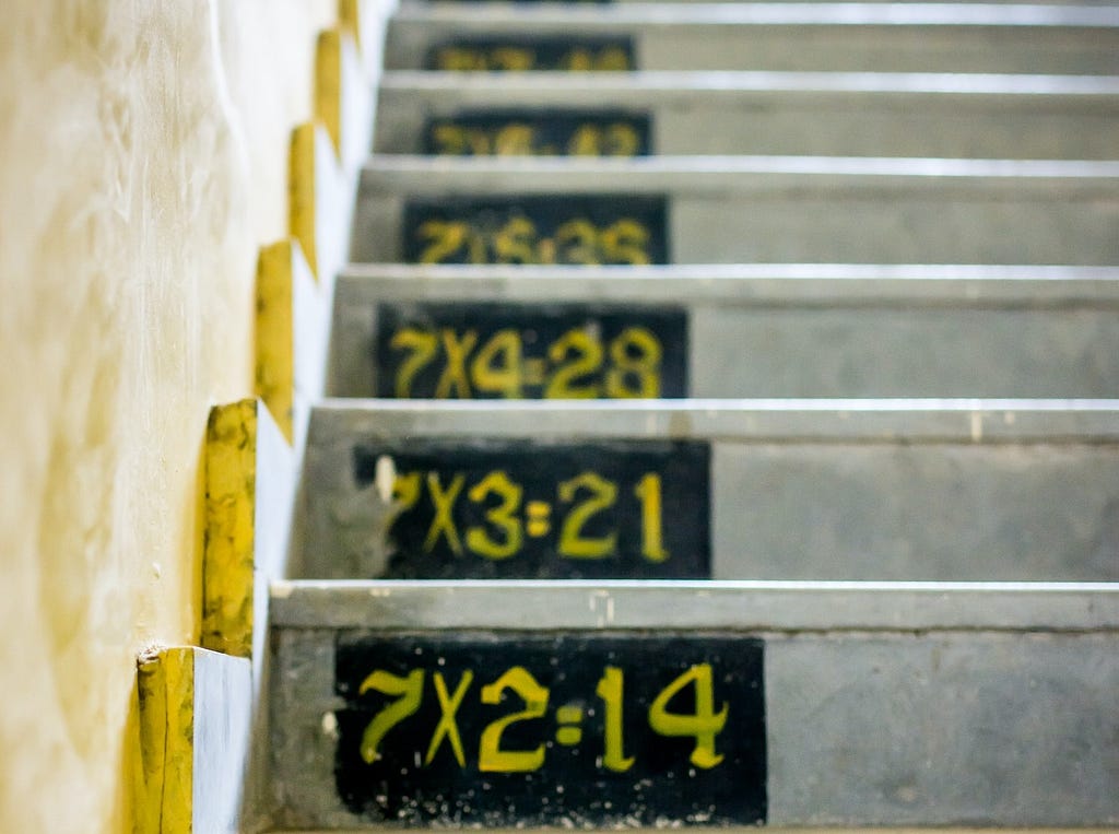 Concrete stairs, probably from a school, with the 7 times tables painted on them, ascending as the stairs go up. The times tables are written in yellow on black chalkboard paint on the left-hand side of each step. The bottom stair shows 7x2=14, then it goes up until the last one you can see is 7x5=35, but it’s clear the times tables continue going. Cropped out of the bottom of the picture is 7x1=7.