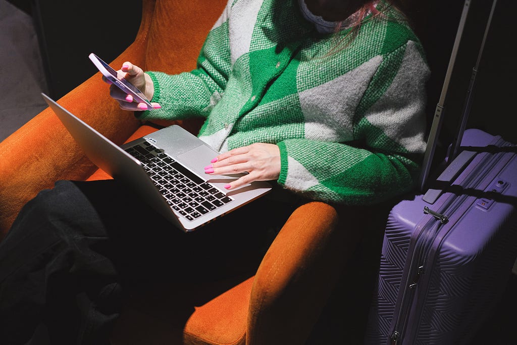 A woman using a smartphone and a laptop while sitting on an armchair.