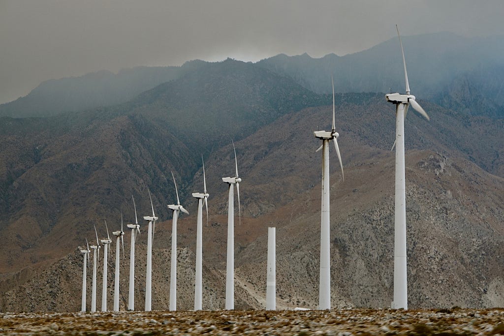 A line of wind turbines stand in front of a mountain range. Smoke hangs in the air.