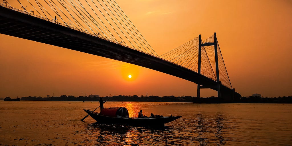 A view of Vidyasagar Setu bridge in Kolkata taken at dusk from one side of the Ganges, with a small barge silhouetted against the river.