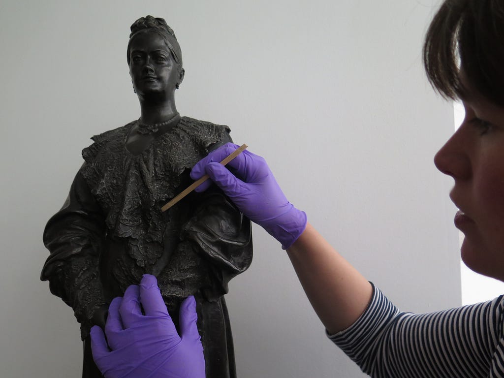 A conservator wearing purple nitrile gloves treats a bronze bust of Enriquetta Rylands with a cotton swab.