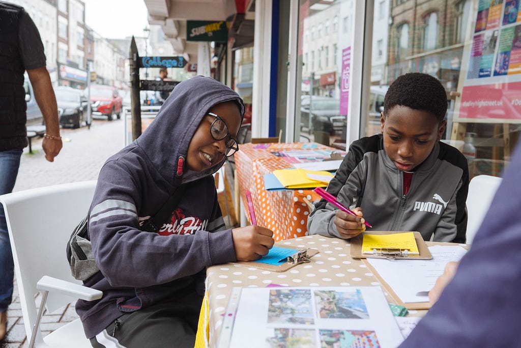 Two young black boys sitting on white chairs at a table on Dudley High Street writing poems. One is wearing a blue/black hoodie with the hood up, writing on bright blue paper. The other is wearing a grey and black sports top, and is writing on bright yellow paper.