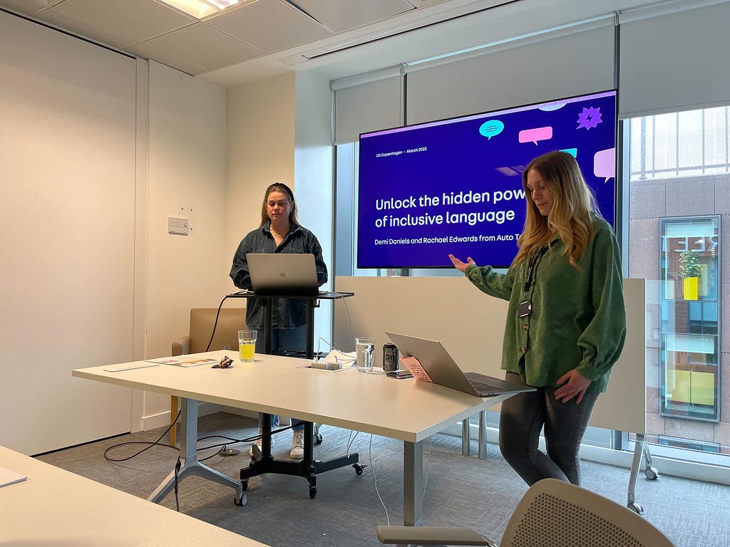 Photograph of Rachael Edwards and Demi Daniels standing in front of a screen showing our title slide from our workshop slide deck at the Auto Trader office. We are about to start our first internal practice run-through for our workshop, which we are presenting to a group of colleagues.