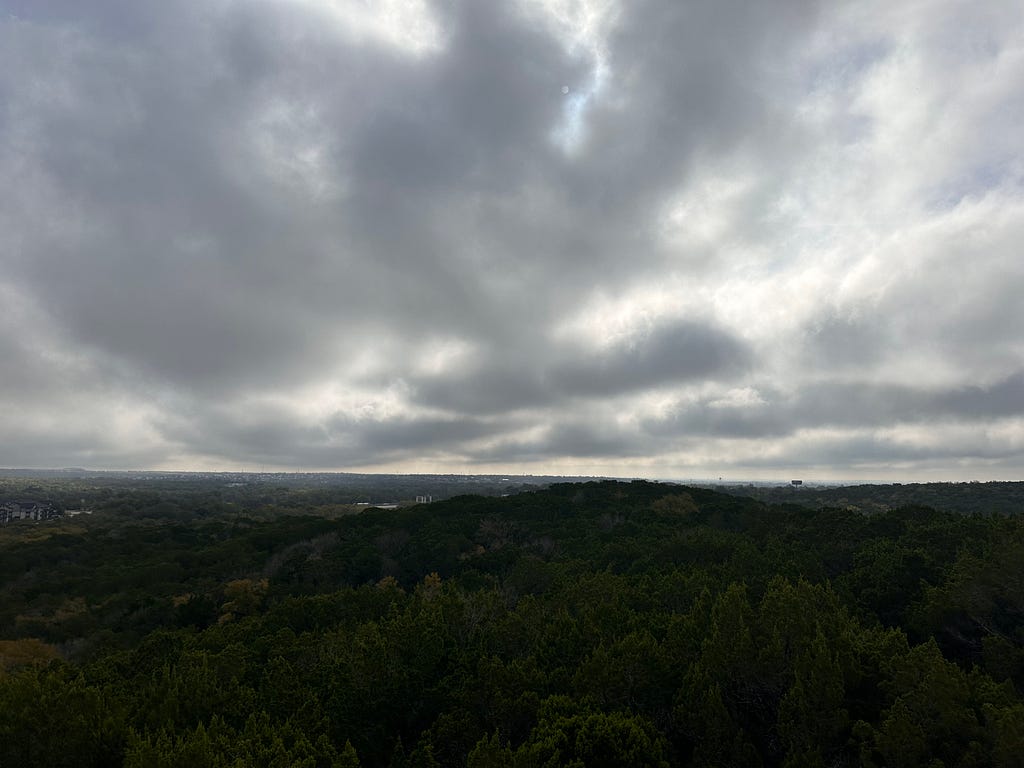 Cloudy sky over a wooded hilly area, with a city in the background.