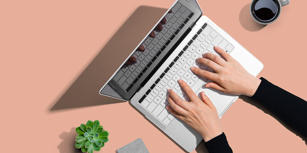 Two hands resting on the keyboard of a laptop, next to a succulent houseplant, a cup of coffee, and a small notebook.