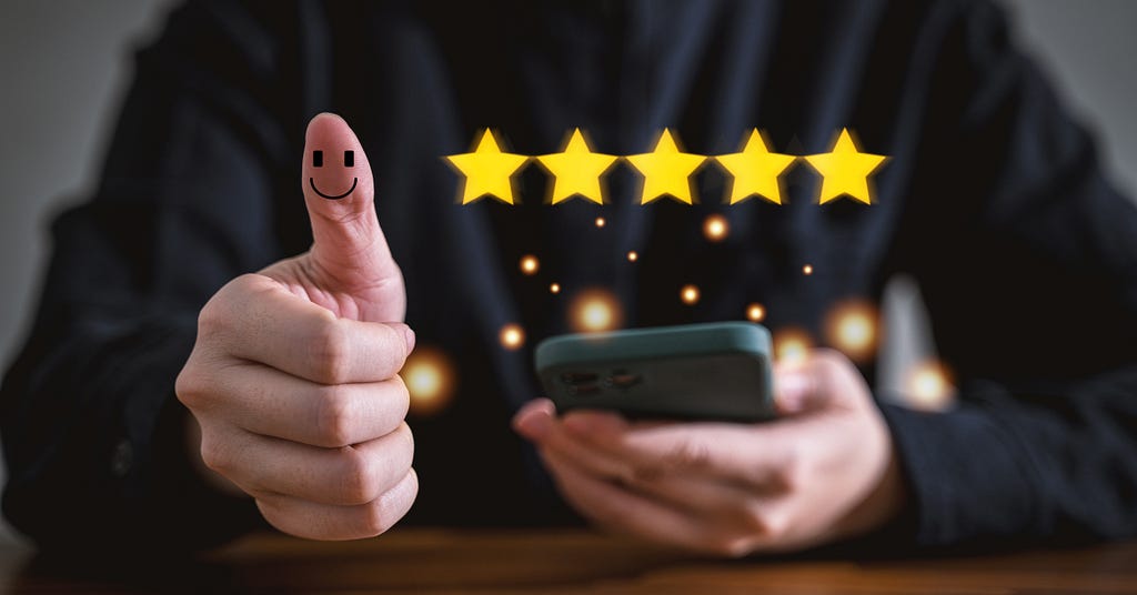 Five yellow stars in front of a person’s black-clad torso with one hand with a raised thumb with a smiley face on it and the other holding a phone.