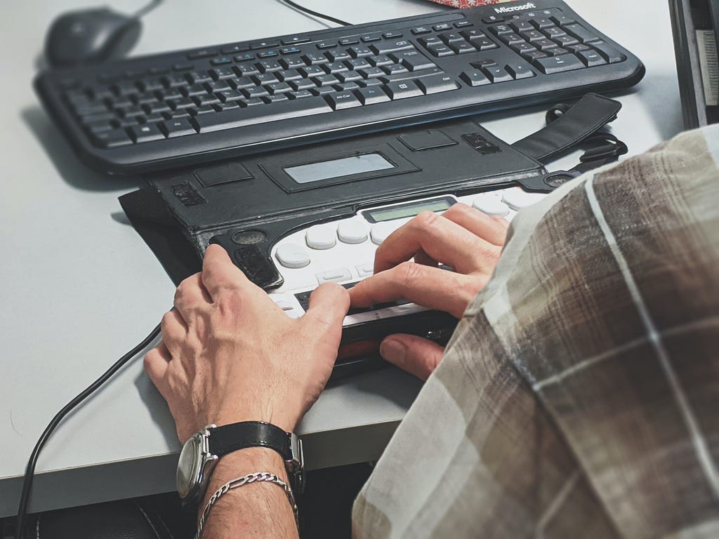 A light-skinned person using an assistive keyboard device to navigate through a website. The image shows a closeup of a person's hand, wearing a watch with a black band, a silver chain bracelet, and a brown and white plaid casual shirt; they are touching their assistive keyboard.