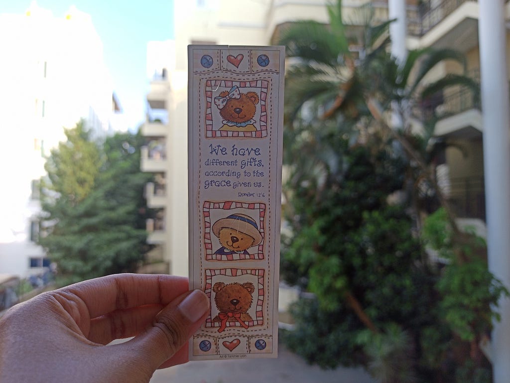 A bookmark with three images of teddies on it is being held up against the backdrop of a tree. The topmost teddy has a bow on its head, the middle one has a hat, and the lower one has a bow on its neck. These words are on the bookmark: “We have different gifts according to the grace given to us.”