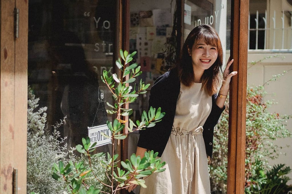 an asian woman wearing an apron who works as a retailer walks out of her store smiling