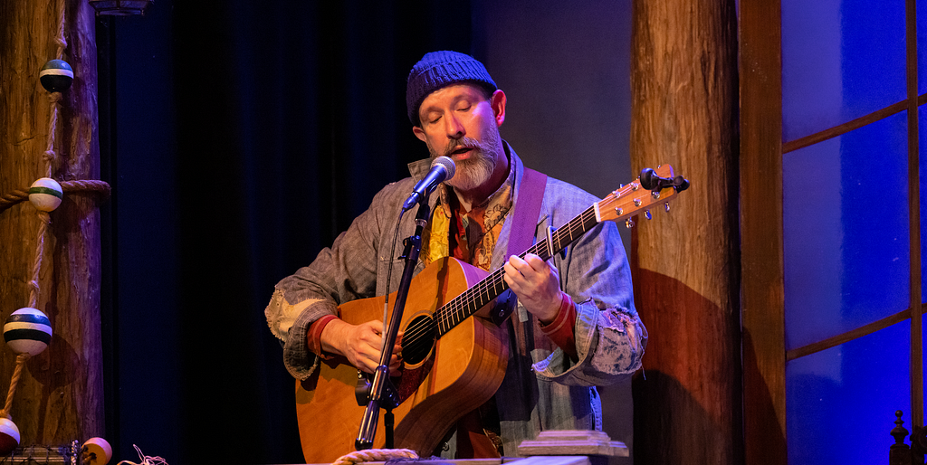 A performer with a beard, in a beanie and denim coat, plays guitar and sings into a microphone.