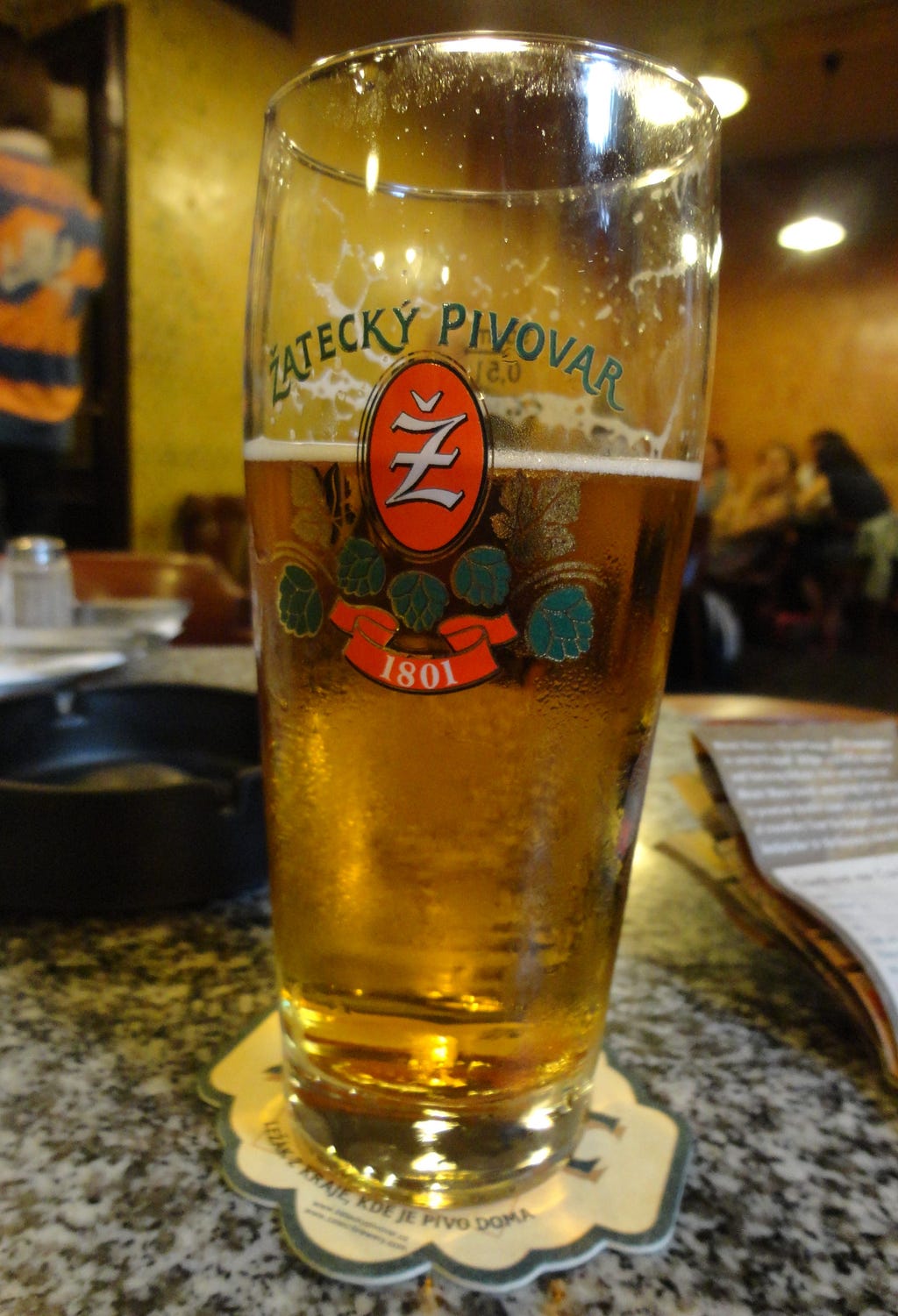 a glass of beer on a table, with the label “Zatecky Pivovar”. An ashtray is also present