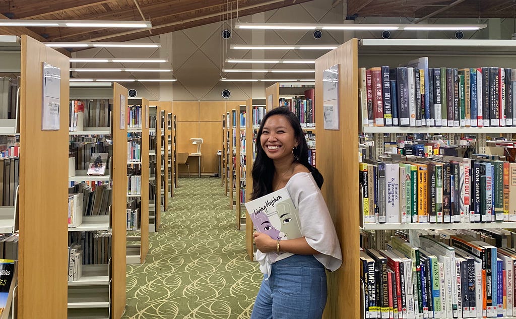 Woman standing against bookshelf amongst library stacks holding Living Hyphen in her arms.