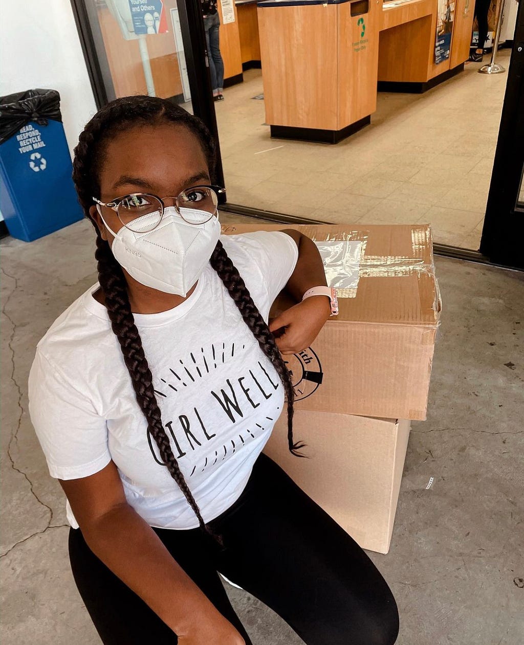 Young girl with box braids and glasses sits in front of stacked boxes. She is wearing a white shirt that says “Girl Well” in black letters.