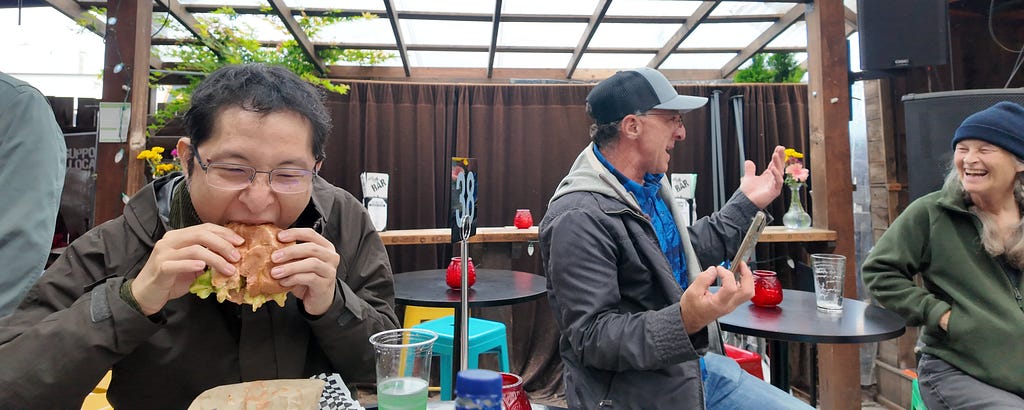 Three people sit around a table at a bar, laughing and eating. One person takes a big bite of a sandwich while the others react in conversation. The space is covered with wood beams, flowers on the tables, and a casual happy hour energy.
