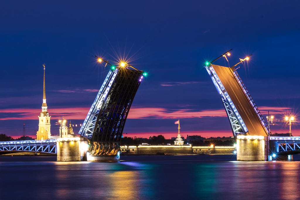 a riverine body of water at sunset with an open bascule bridge. there is a brightly lit building with a spire to the left in the background, and a building in the center farther back with an American flag flying above it.