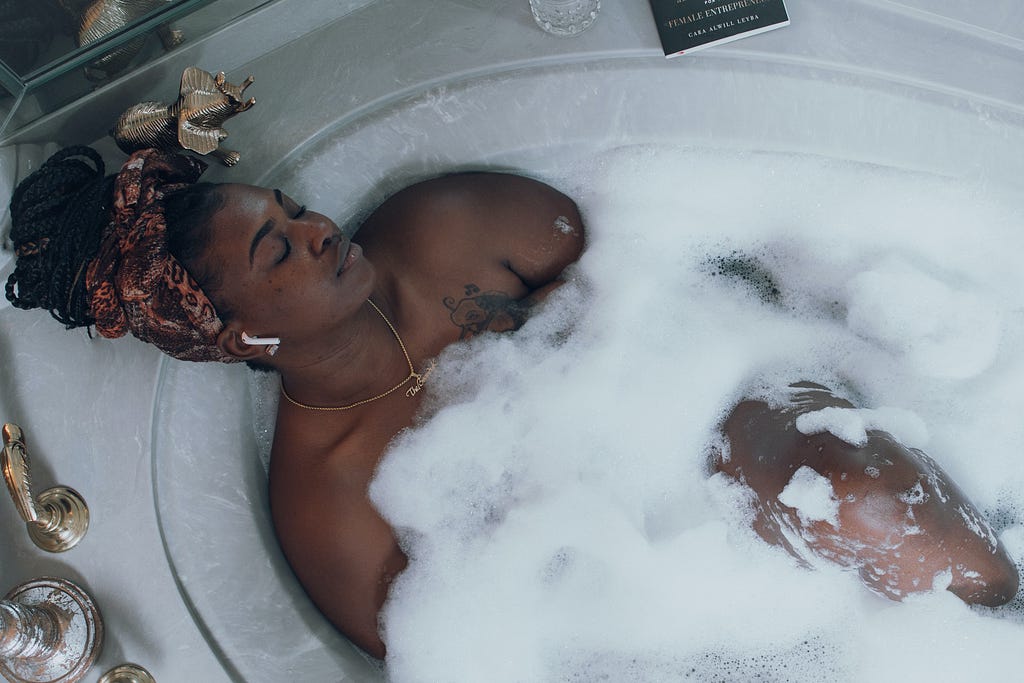 A Black person laying on their back in a bubble bath in a stone bath tub. They’re wearing a patterned headwrap to keep their braids out of the water. They have PERFECT eyebrows, wow, and have their eyes closed, facing away from the camera. They have Airpods in, and a necklace around their neck resting on their chest. They also have a tattoo on the left side of their chest.