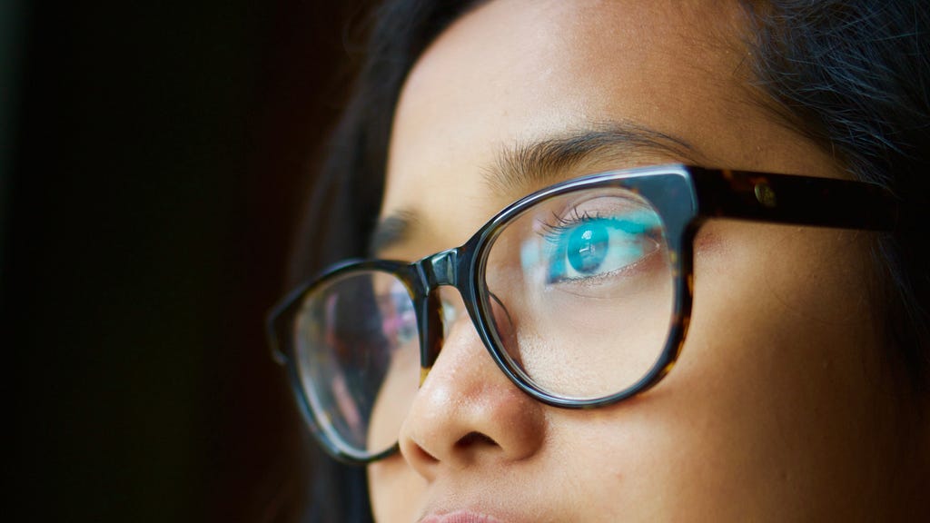 The upper side of an asian woman’s face. She is staring at a monitor out of frame. The monitor is reflected  on her glass lenses.