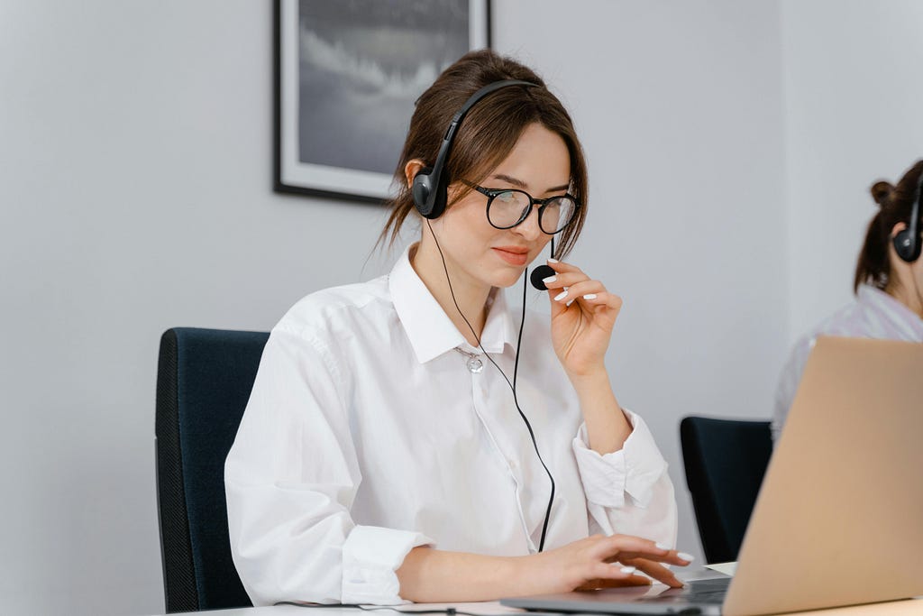 A woman customer service agent multitasking with headphones and a headset while working on a laptop.