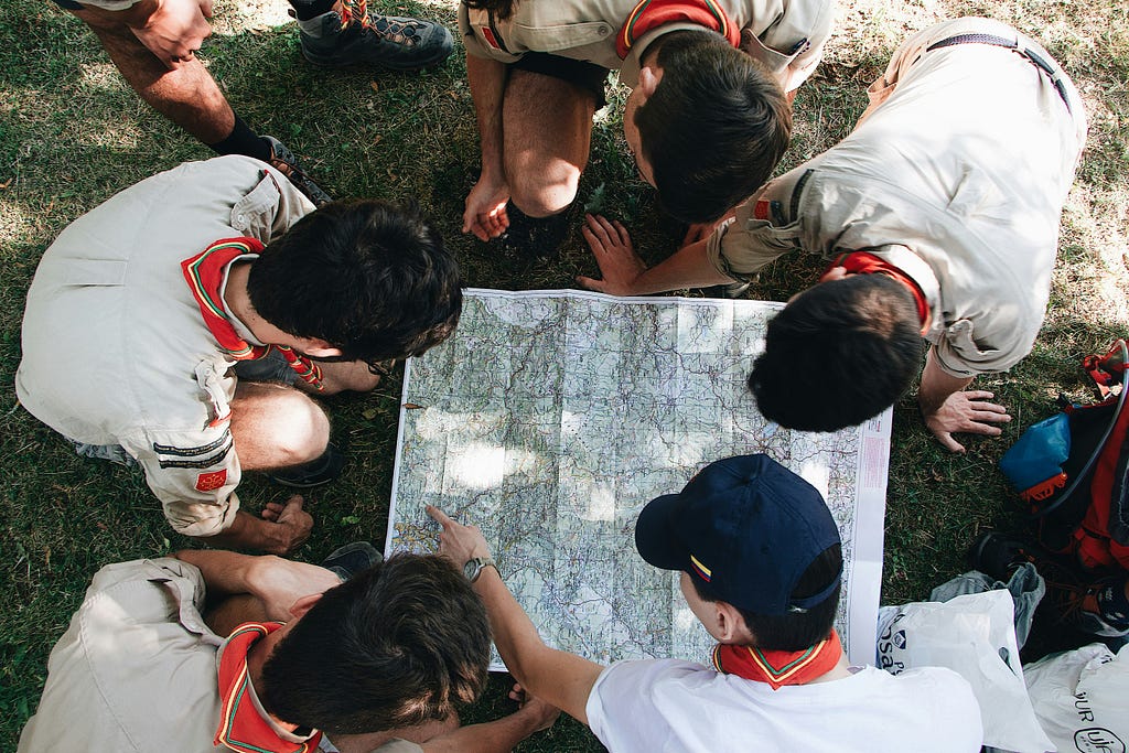 Exploring! A group of uniformed boy(?) scouts outdoors, crouched down around a map spread out on the grass and pointing out a location.