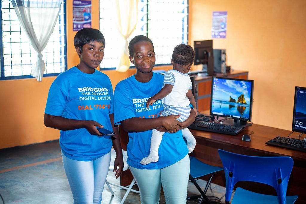 Two women — one of them holding a mobile phone and the other cradling a small child in her arms — stand next to a table with computers while wearing T-shirts that say “Bridging the gender digital divide.”