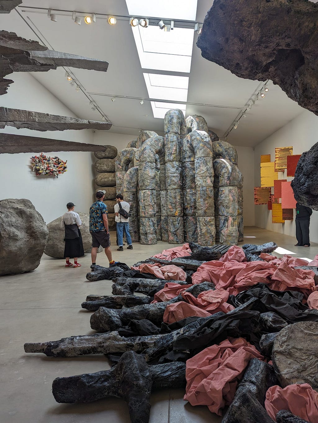 A large gallery space filled with several sculptures by Phyllida Barlow. On the floor in the foreground is what looks like a pile of black and pink fabrics; to the left are what look like 2 big boulders; at the back of the room are huge pipe-like structures reaching the ceiling; on the right wall are yellow, pink and red shapes fixed to the wall. 3 people are wandering and looking around.