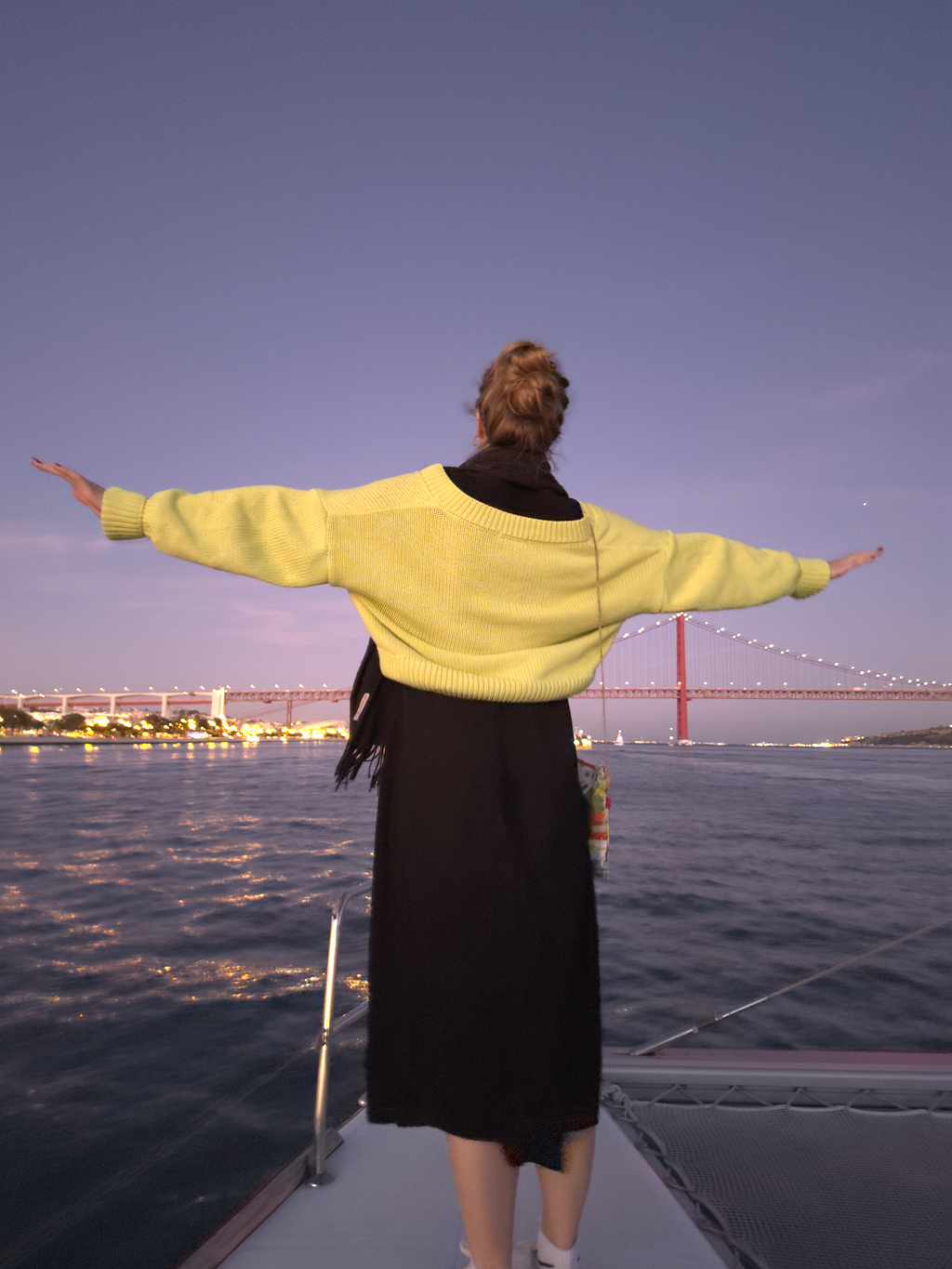 girl standing on the edge of a boat looking at the city of Lisbon in the background and having her hands in the air