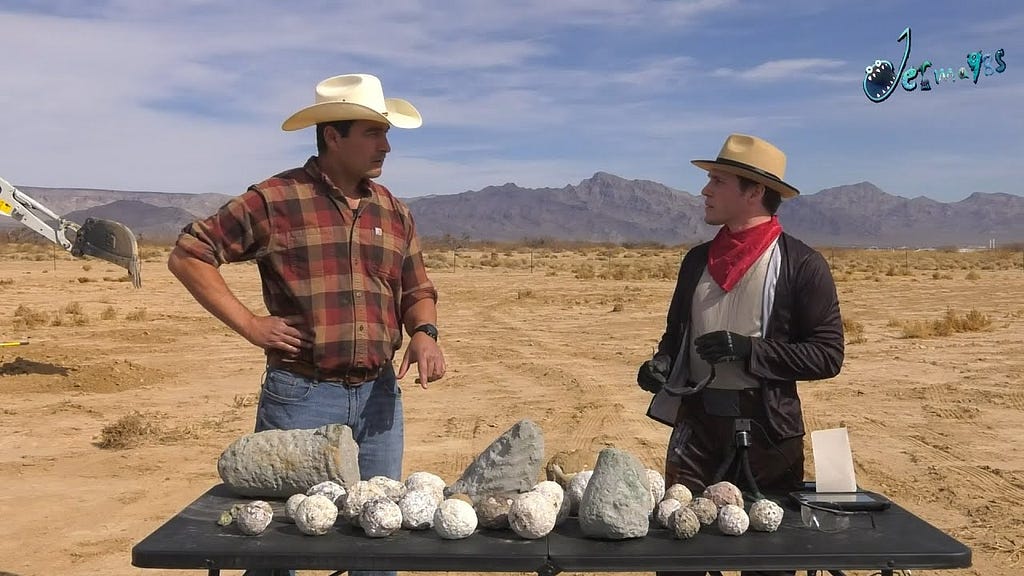 Jerma985 dressed as a cowboy with archaeologist Josh, at a table with rocks on it in front of a dusty/arid landscape