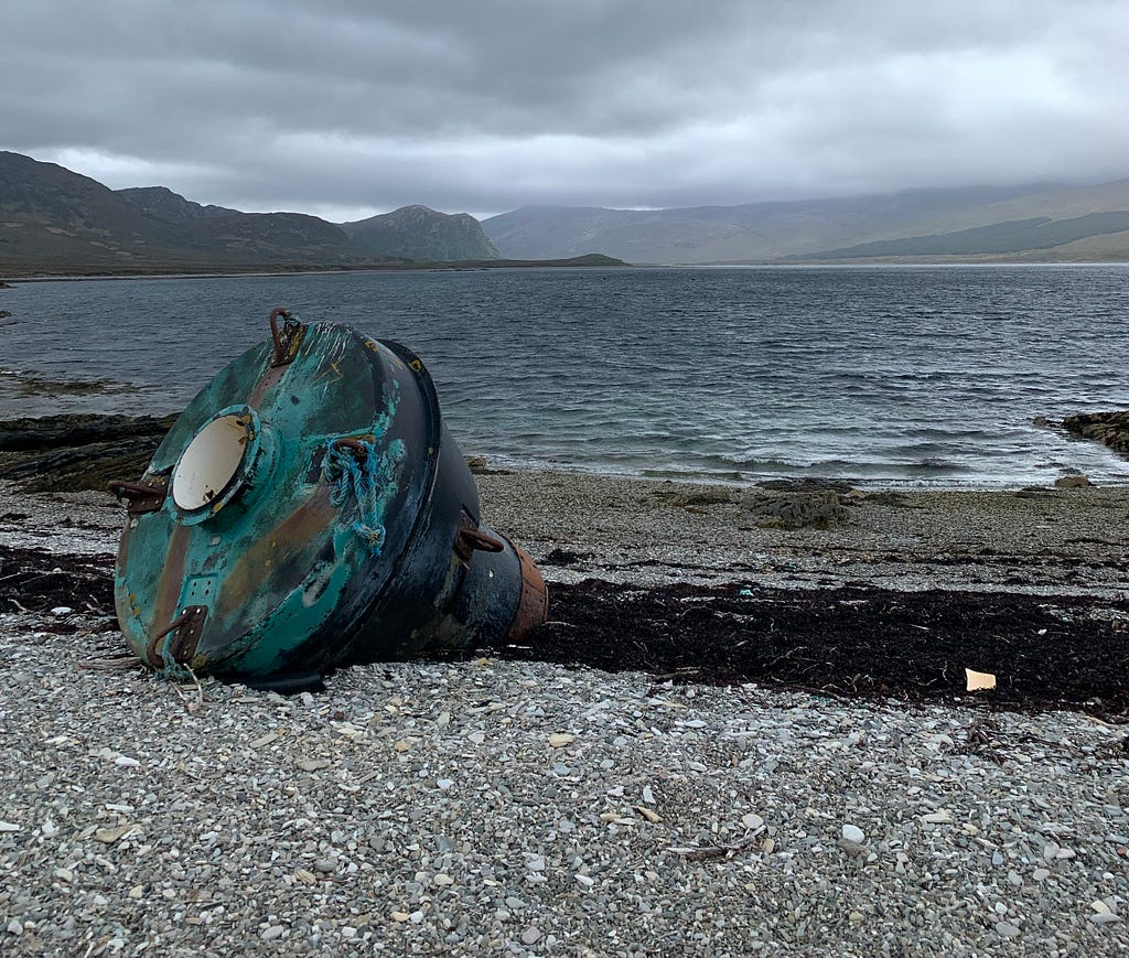 A weathered sea buoy washed up on the shores of a Scottish sea loch under grey skies
