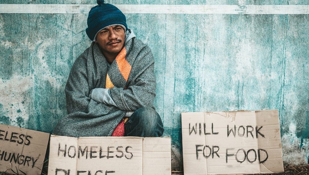 Homeless man with back against a wall and supplication signs — one says “WILL WORK FOR FOOD”.