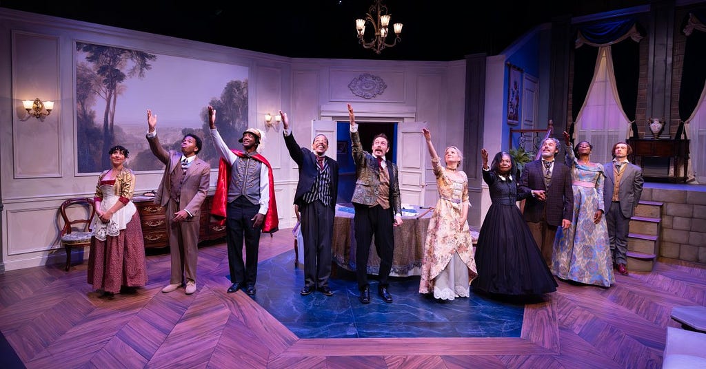 Ten actors stand in a line on stage for a curtain call, dressed in full period costume and raising their right arms to salute the production’s stage manager.