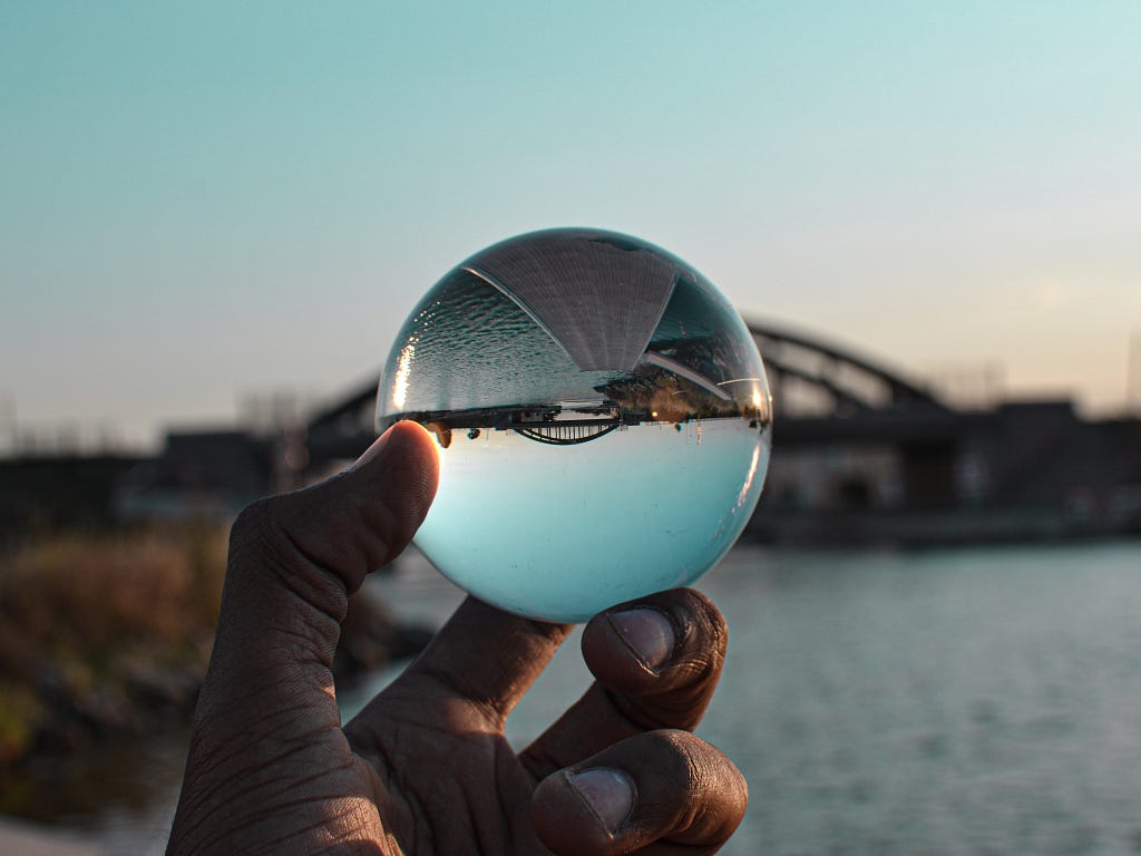 A hand holds up a glass sphere, showing an upside down perspective of a bridge over water.