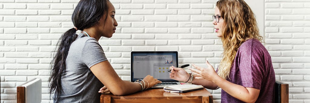 Image of a black woman and white woman talking. White woman is taking notes while both look at a laptop screen.
