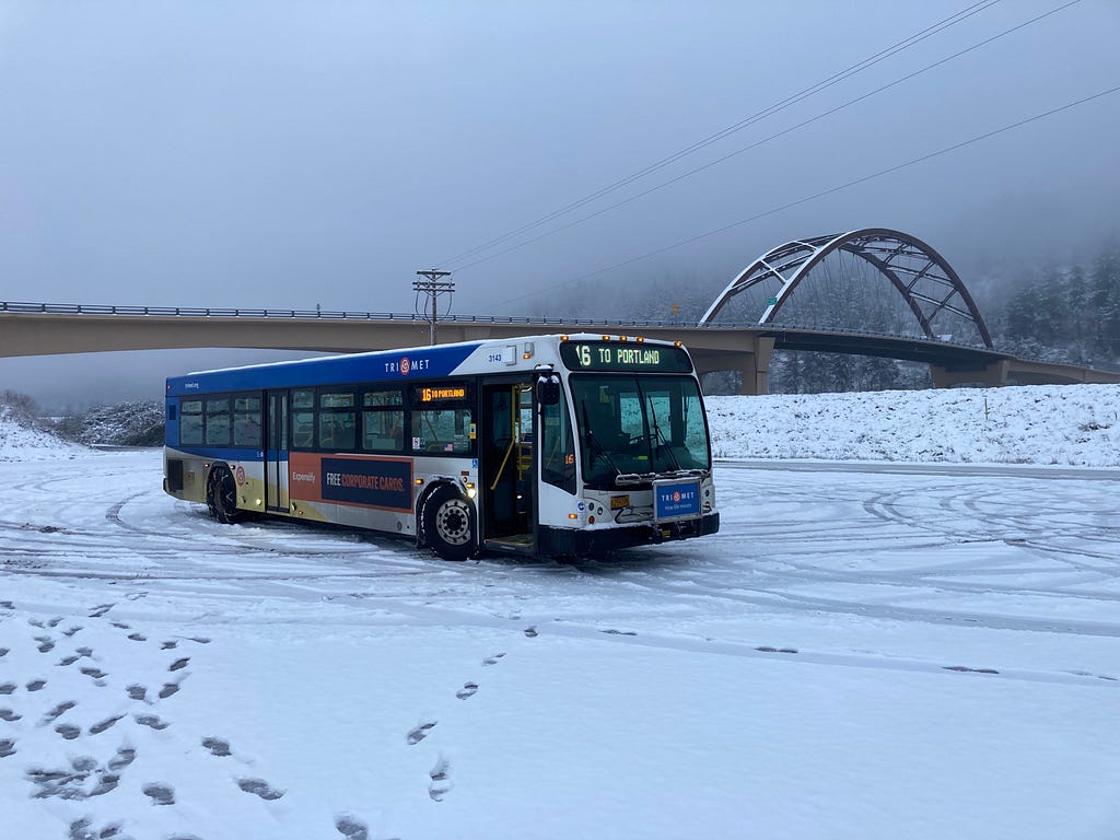 A bus parked in the snow with footprints leading from it with a view of the Sauvies Island Bridge in the background.