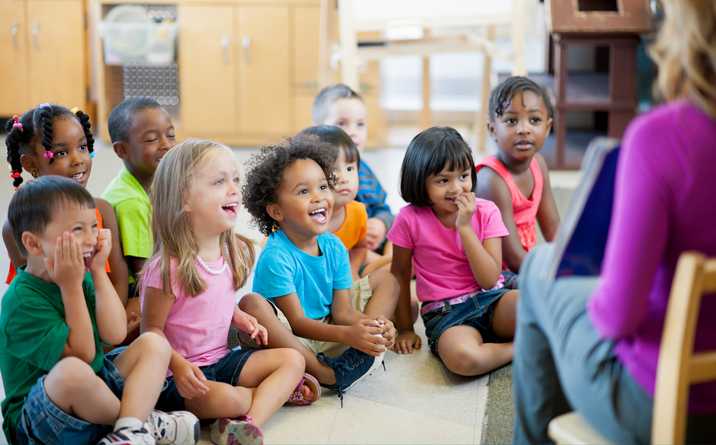 Kindergarteners sit on the floor in their classroom listening to their teacher reading a story.