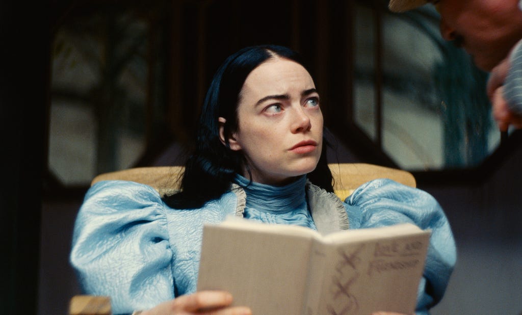Bella Baxter (Emma Stone), a white woman with black hair and blue eyes, wears a Victorian gown and reads Love and Friendship.