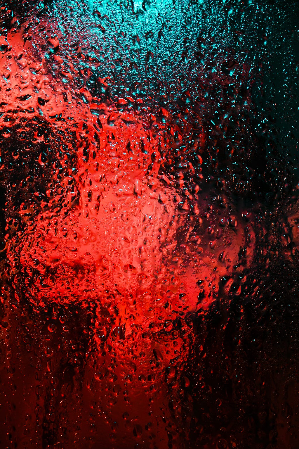 A window filled with droplets of water and red and turquoise light emanating from the other side.