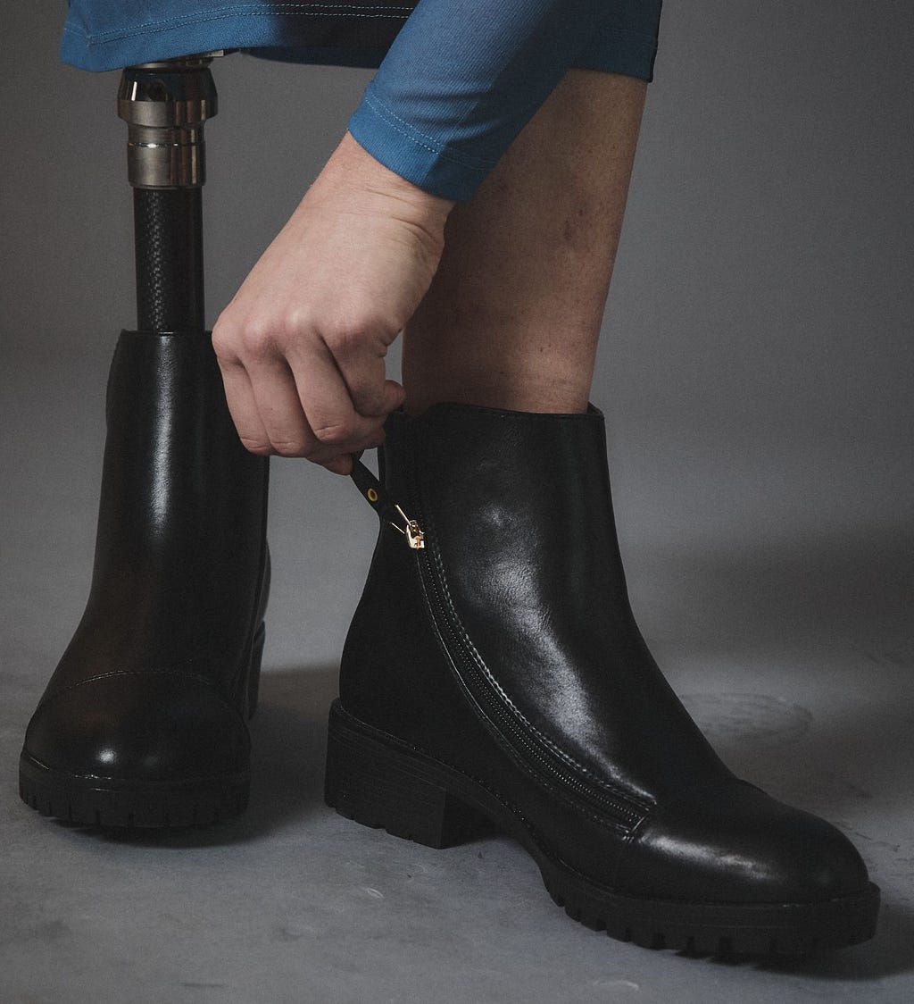 A person’s hand zipping up a pair of Adaptive black leather boots, worn by someone with one prosthetic leg.