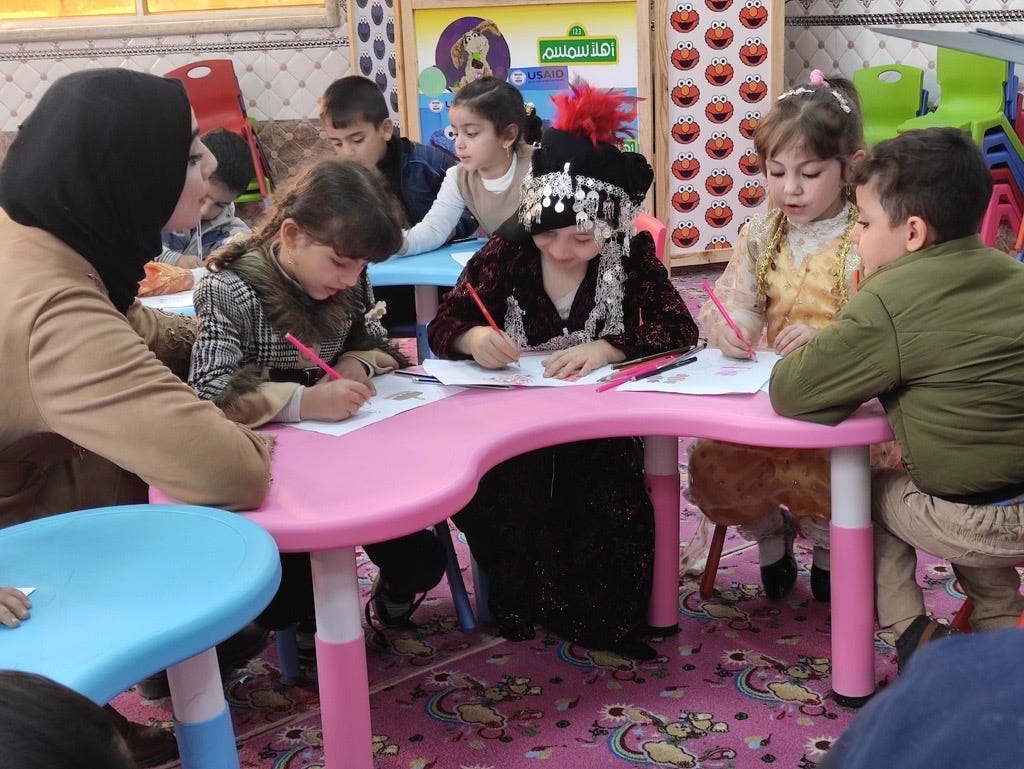 A woman observes three girls and one boy drawing while wearing traditional Iraqi dress.