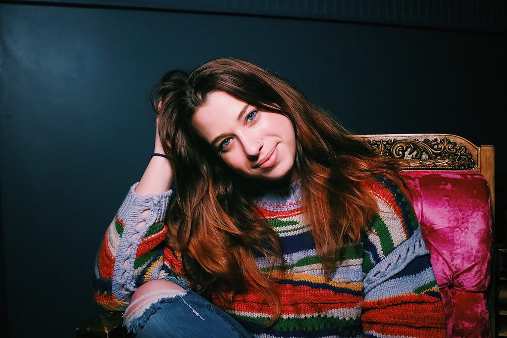 Ariel Elias, white female, sits on a wooden chair with pink cushioning in front of a dark green wall. She is in a multi-colored sweater and torn jeans. Elias sits with her hand on her head, mixed into her hair, and her head gently tilted to the side. Elias has long brown hair.