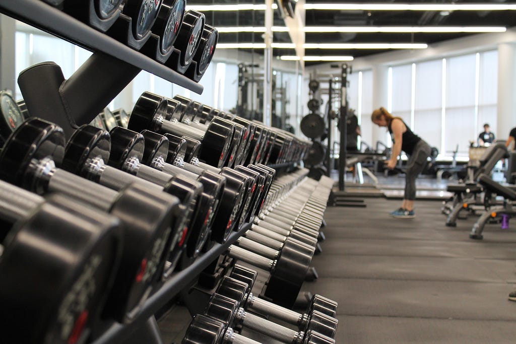 A photograph of a gym. In the foreground is a closeup of a rack of barbells, and in the background is a woman lifting weights.