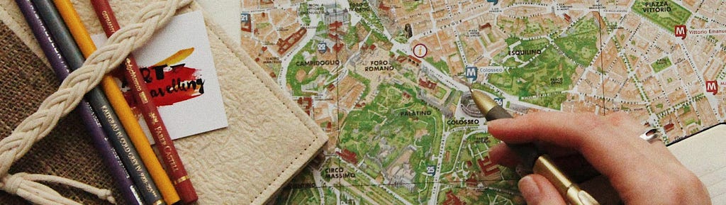 A hand holding a pen hovers over an illustrated map of a city with points of interests and areas labeled. The left side of the photograph shows a notebook and colored pencils.