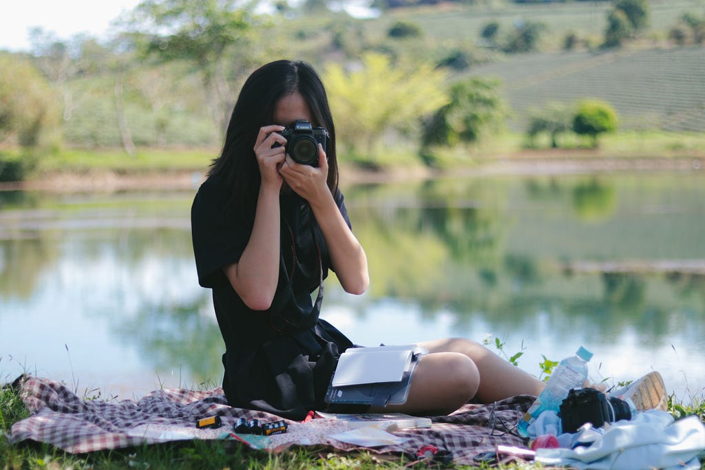 A woman sitting on a blanket near a lake practices the hobby of photography by taking a picture with the camera facing the reader. There is a notebook in her lap and a water bottle my her feet. The background is hilly with trees.