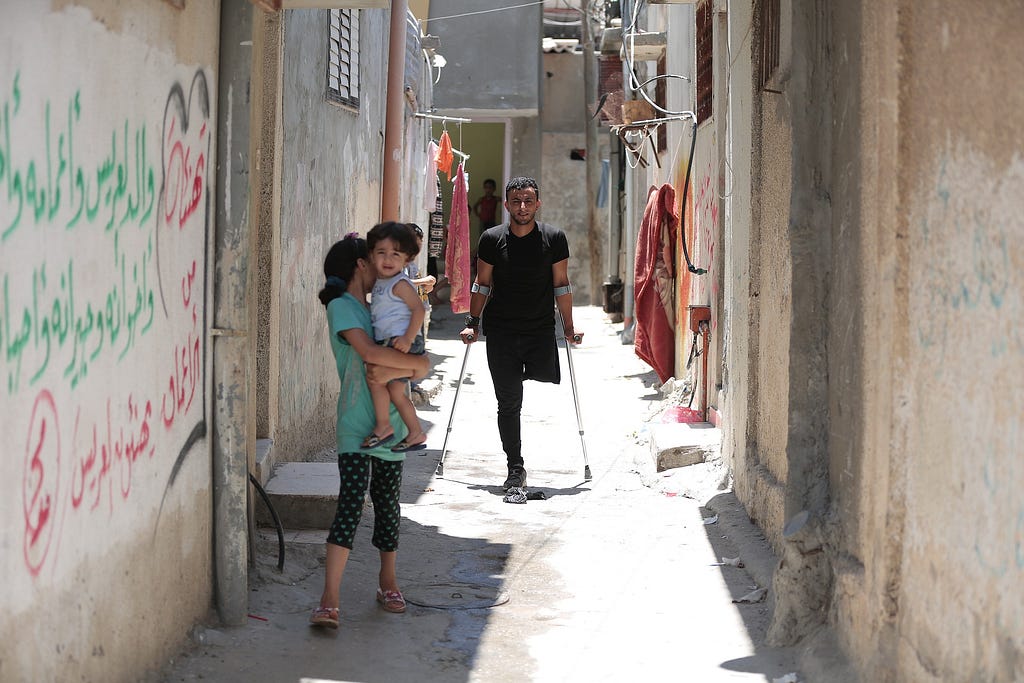 Narrow alley between buildings in the Gaza Strip. A girl holds a smaller child in her arms. In the background, a man on crutches is missing a leg.
