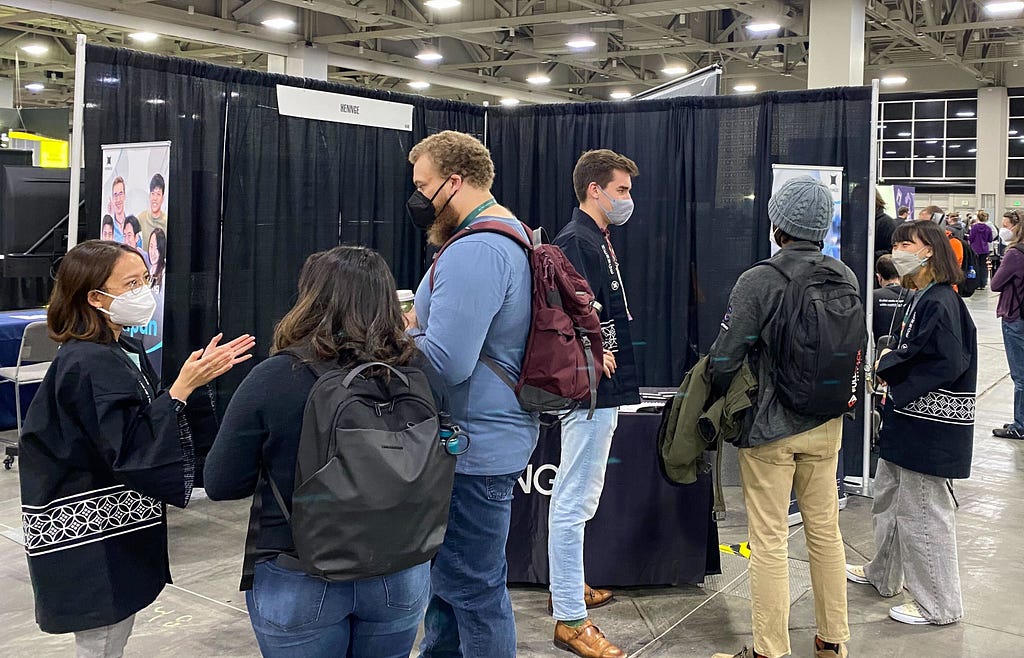 A photo of the booth during the day. The HENNGE people are chatting with attendees.