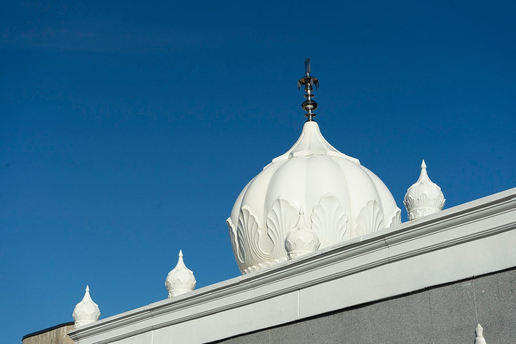 White domes and a silver spire contrast against a blue sky, just outside the Sikh Temple