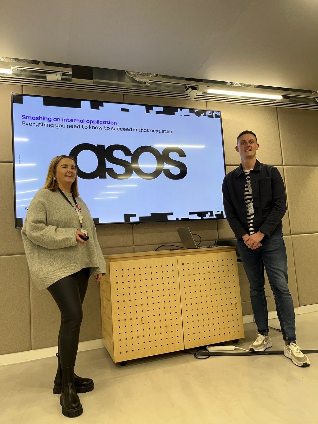 Two ASOSers, Lauren and James, standing in front of a presentation screen that reads ‘Smashing an internal application. Everything you need to know to succeed in that next step’.