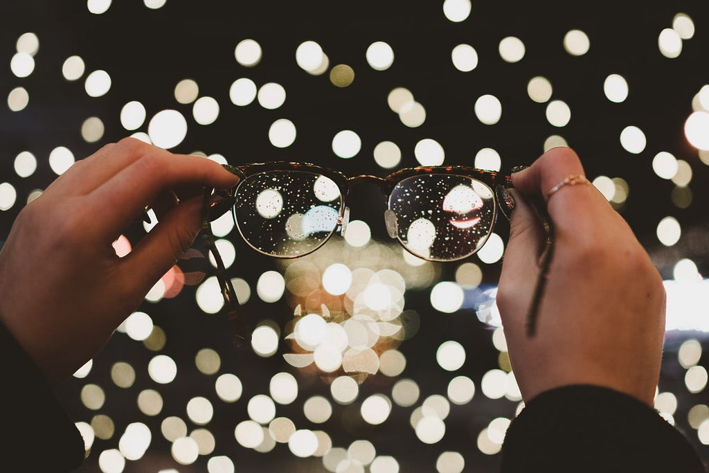 A pair of wet glasses held by a pair of hands in the rain.