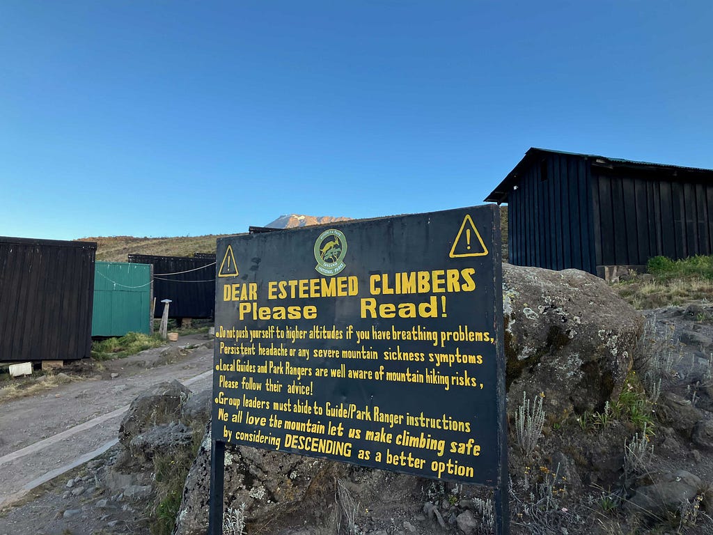 A sign with black background and gold text warning mountain climbers not to continue to higher elevations if they have breathing problems, headache, or other symptoms of altitude sickness.