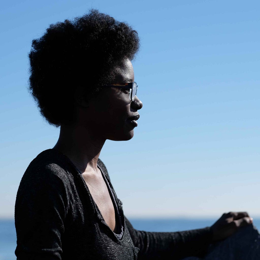 Simone is Black non-binary person pictured at Beach 44 in Far Rockaway, Queens, New York. She sits on and against a rock, resting her left arm and hand on another rock to her left. She is wearing a black, wide neck, long-sleeved shirt. Simone has black hair and is wearing it in a short afro, she also has glasses on. She is looking into the distance. The upper half of the background is a light blue sky that merges into a blurry blue color closer to the bottom of the photo, which is the ocean