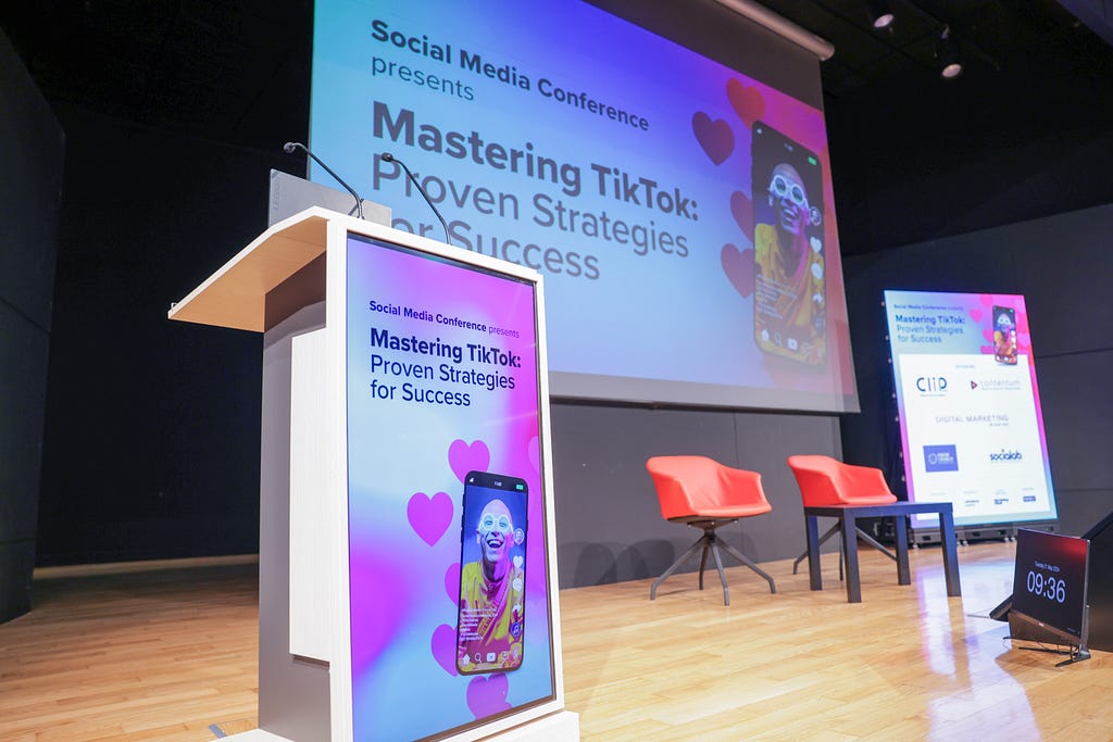 The image depicts a conference stage setup for a presentation titled “Mastering TikTok: Proven Strategies for Success.” The stage features a podium with a screen displaying the presentation title, two red chairs, and a small table. Behind the podium, a large screen also shows the same title along with a vibrant image of a person on a TikTok screen, smiling and wearing round glasses.
